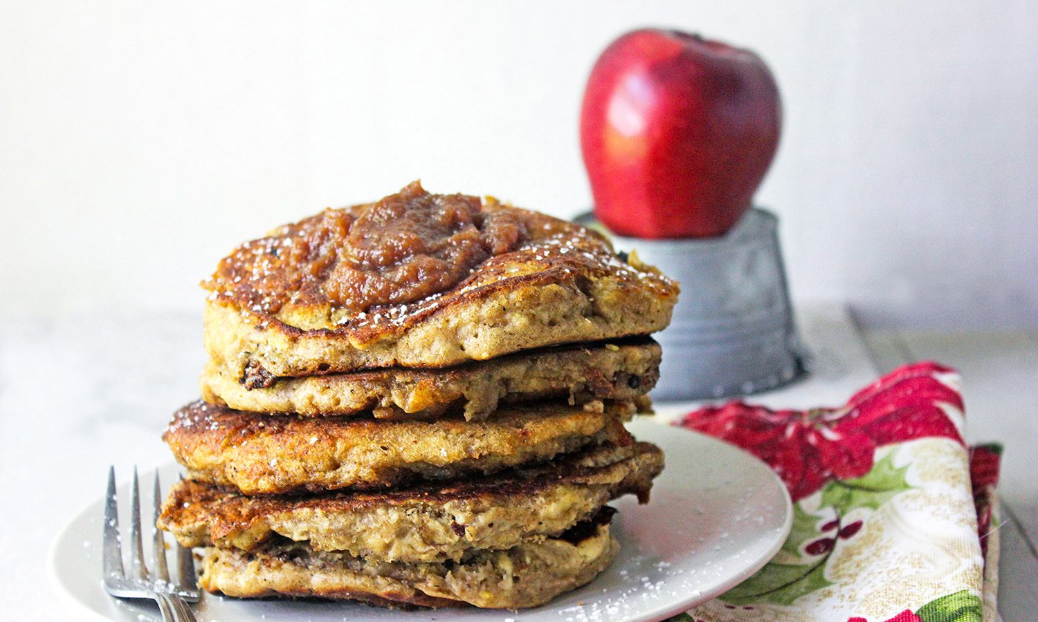 Best Apple Butter Pancakes stacked high and dripping with apple butter plated, sitting on a vintage red tablecloth with a fresh apple in the background.