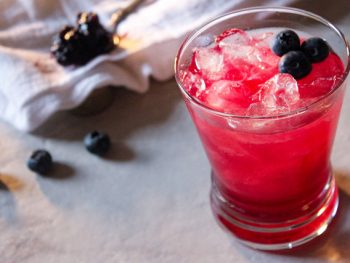 Pinkish hued blueberry bourbon cocktail in a rocks class with fresh blueberry garnish and kitchen towel and spoon of jam in background