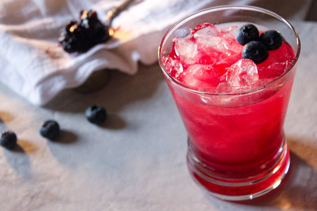 Pinkish hued blueberry bourbon cocktail in a rocks class with fresh blueberry garnish and kitchen towel and spoon of jam in background