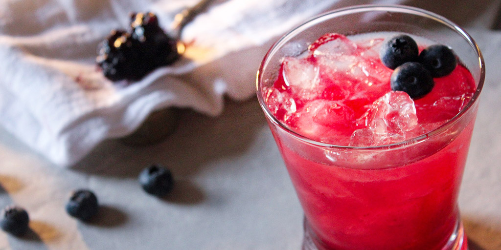 blueberry jam cocktail in rocks glass garnished with fresh blueberries sitting on kitchen counter