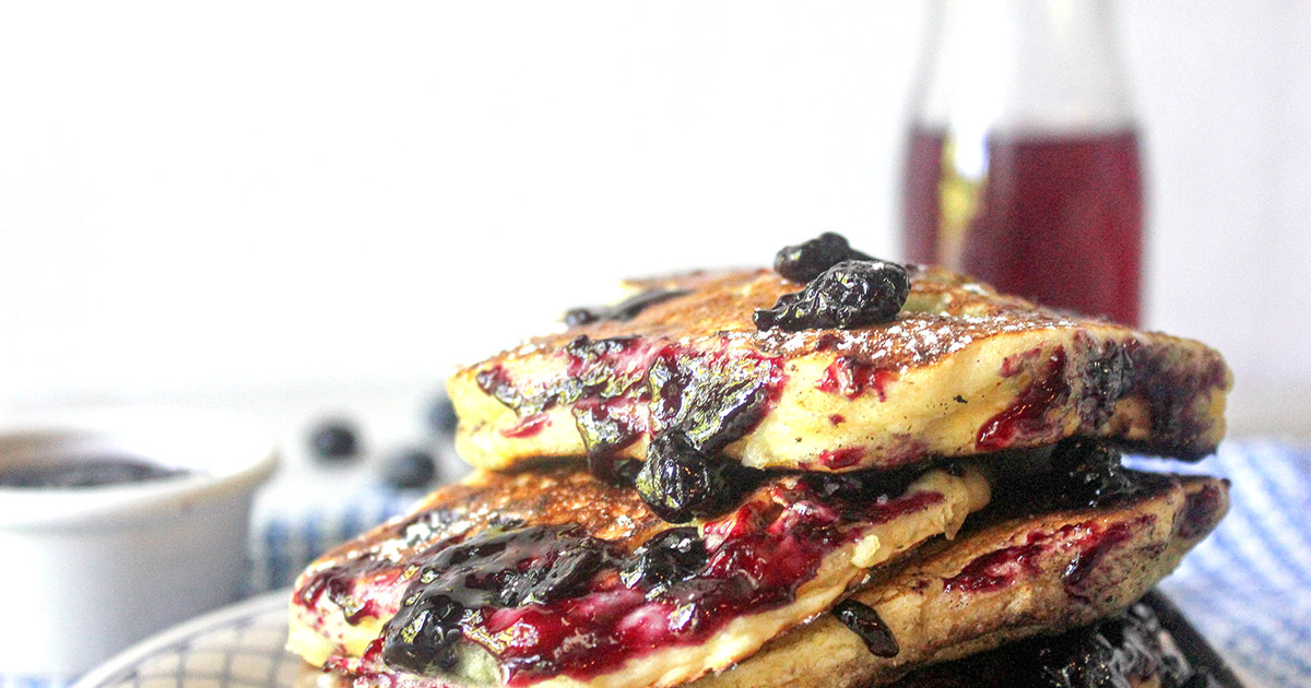 Blueberry Pancakes casually stacked with blueberry jam oozine over the top and blueberry jam syrup milk jug in background
