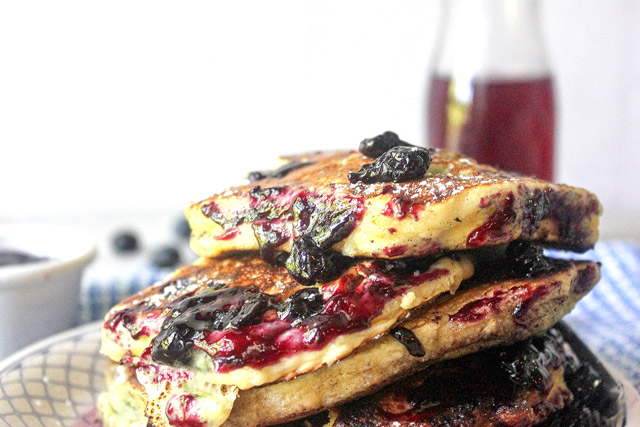 Staggered stack of blueberry pancakes with blueberries and blueberry jam syrup dripping down sides