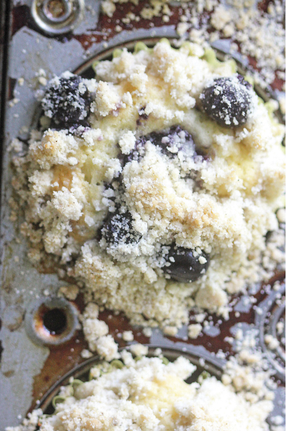Cardamom blueberry muffin in tin showing crumble top and blueberries.