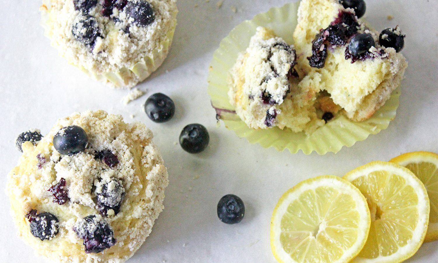 Cardamom blueberry and lemon muffins with a crumbly top spread on white marble with blueblerries and lemon rounds.
