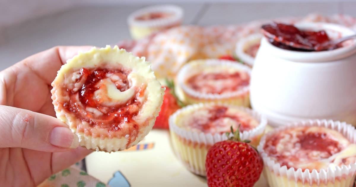 Hand with individual cheesecake with strawberry jam swirls - strawberries and desserts in background