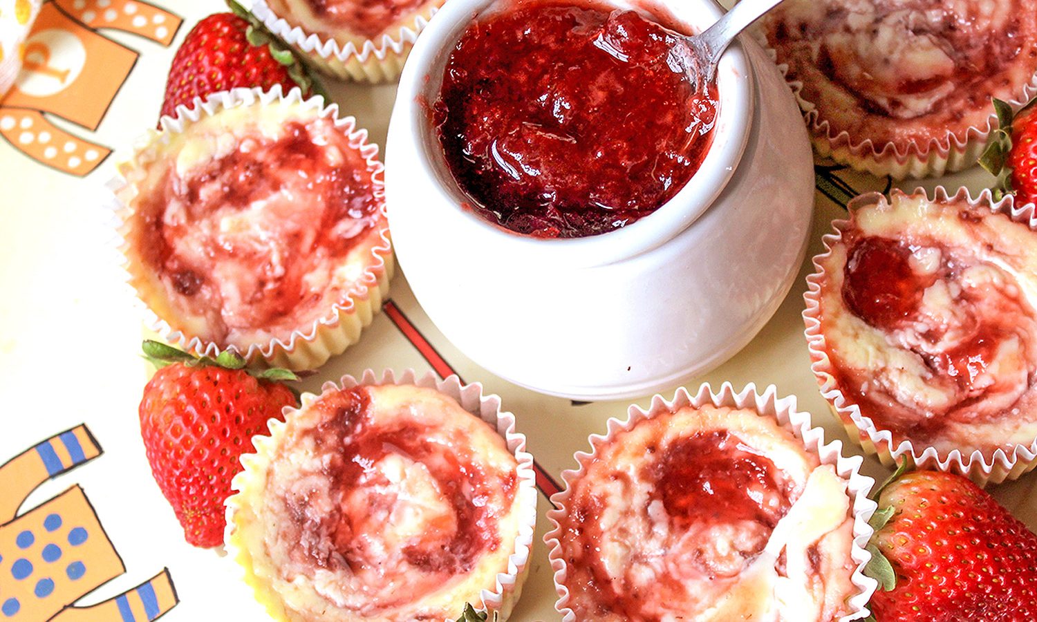 Individual cheesecakes with strawberry jam swirls and a jam pot filled with strawberry jam