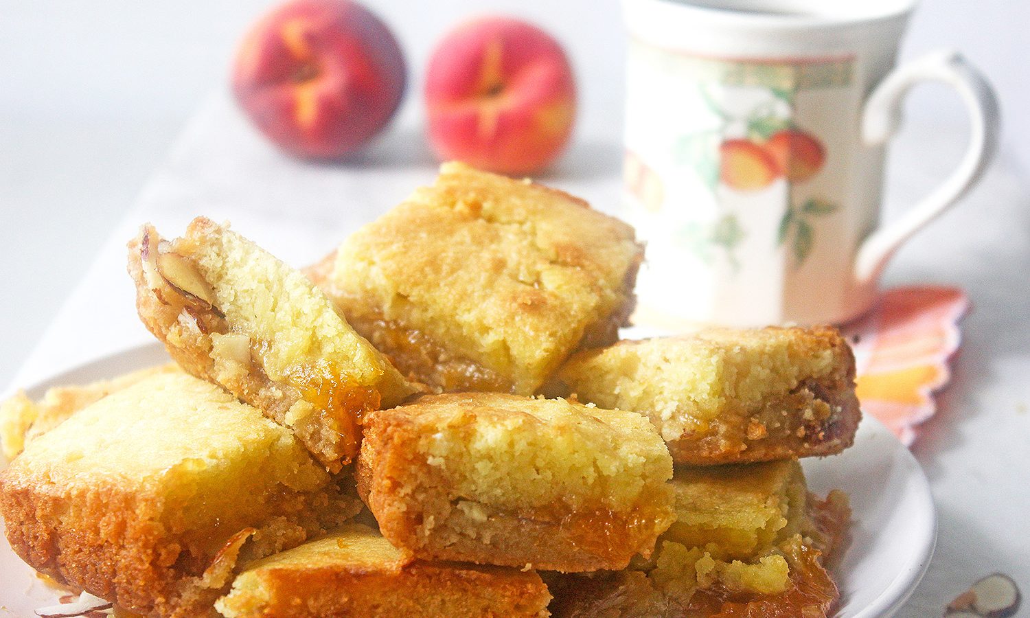 Peach cobbler jam bars stacked on plate with fresh peaches and a peach motif teacup in the background.
