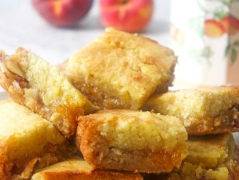 Plate full of casually stacked peach cobbler jam bars with a peach themed tea cup and fresh peaches in the background