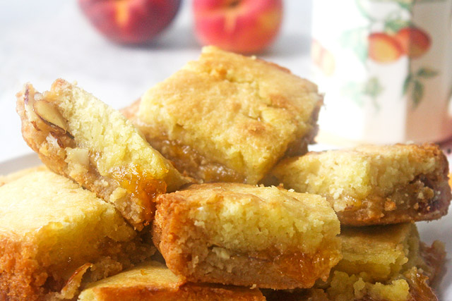 Plate full of casually stacked peach cobbler jam bars with a peach themed tea cup and fresh peaches in the background
