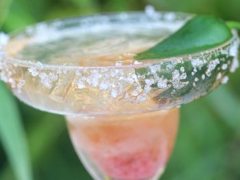 Strawberry jalepeno jam margarita in traditional glass rimmed with salt, garnished with jalepeno. The cocktail is a peachy red.