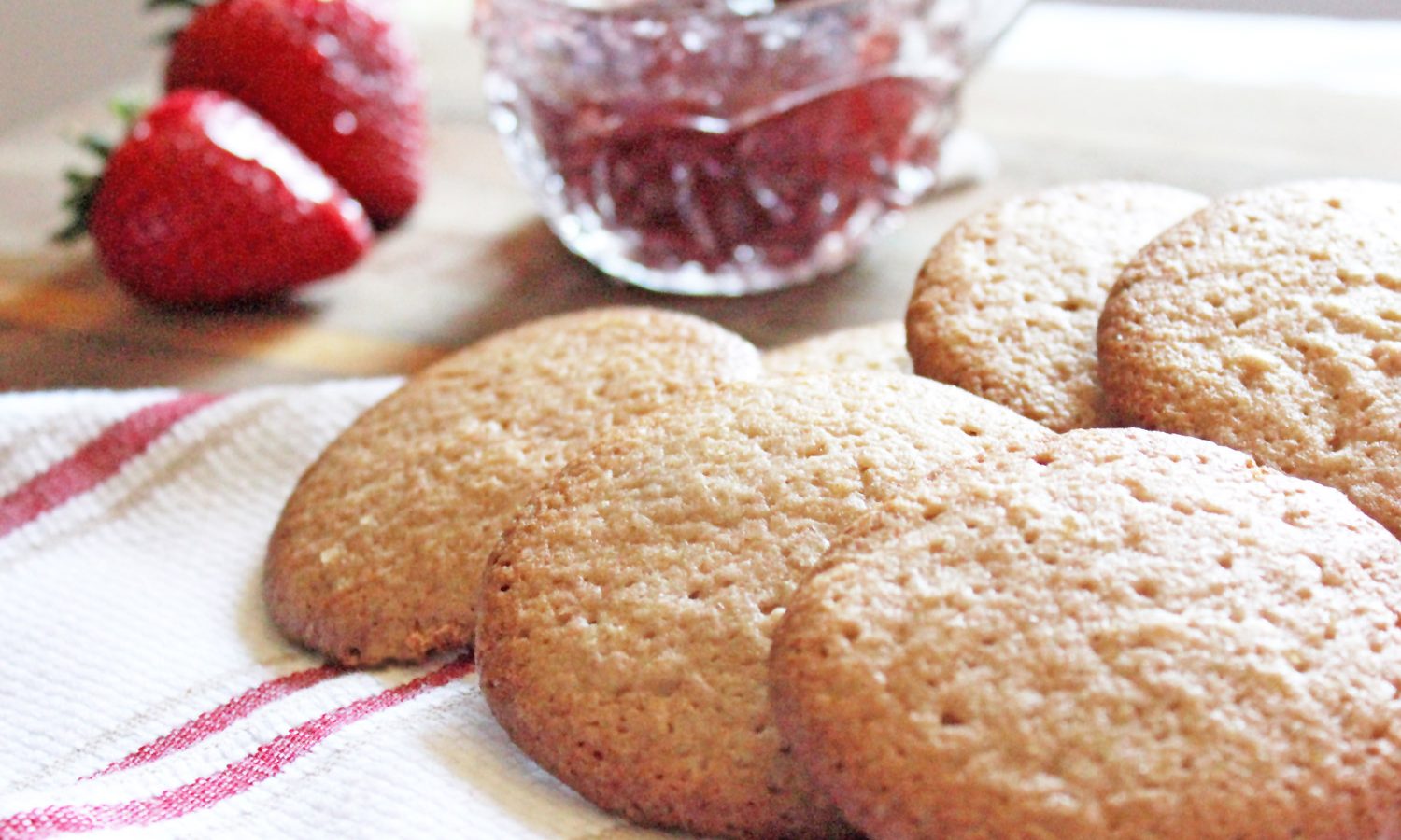 Strawberry jam sugar cookies spread on red and white towel with fresh strawberries and a crystal bowl of strawberry jam