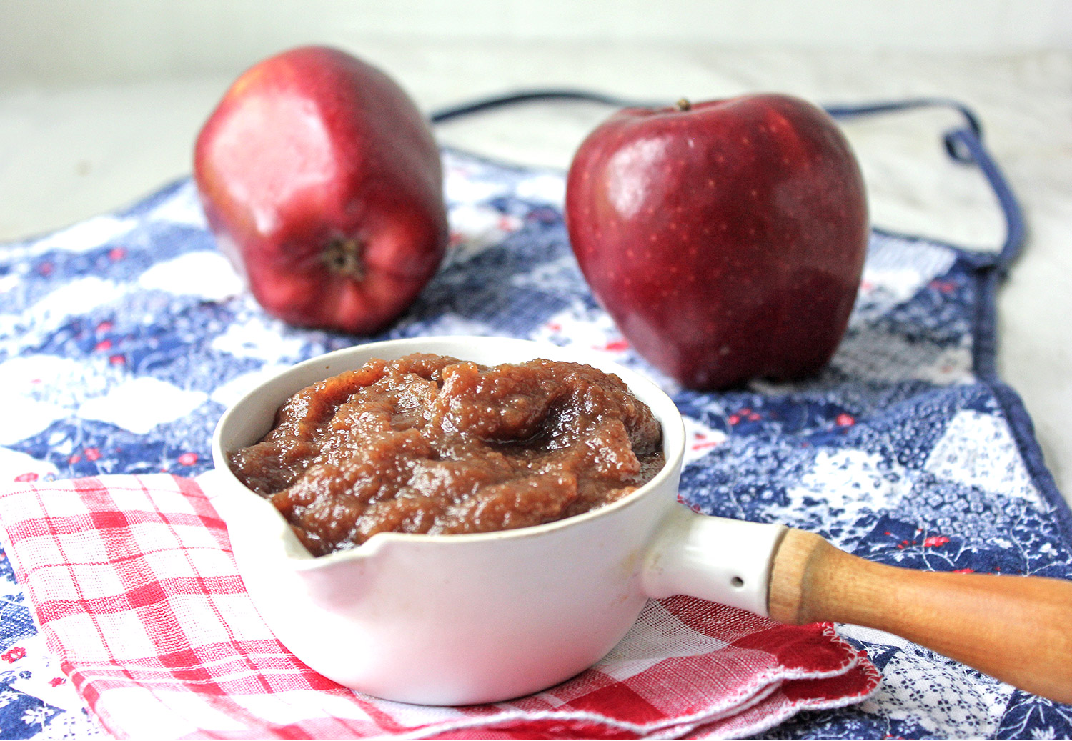 Apple butter in a small enamel pouring cup resting upon an apron with apples in the background.