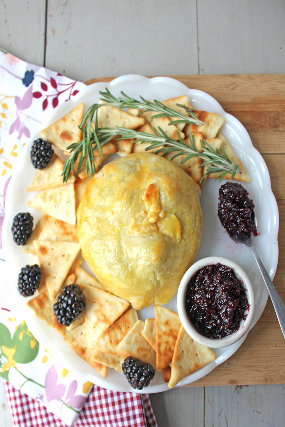 Uncut baked brie and berries with jam on platter garnished with fresh blackberries, rosemary and blackberry bourbon jam.