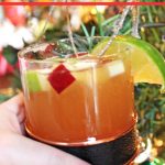 Hand holding bourbon apple cider cocktail in front of Christmas tree.