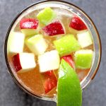 Top view of bourbon and apple cider cocktail showing red and green cubed apples and lime wedge garnish.