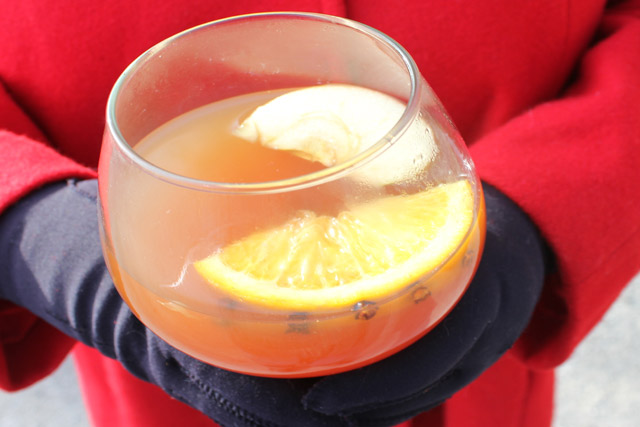 Snifter bowl glass with apple cider bourbon wassai held in gloved hands next to a red wool coat